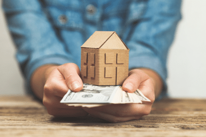 A person holding cash in their hand with a cardboard house on top of the cash representing the cash at close you'll likely receive when selling your vacation rental business